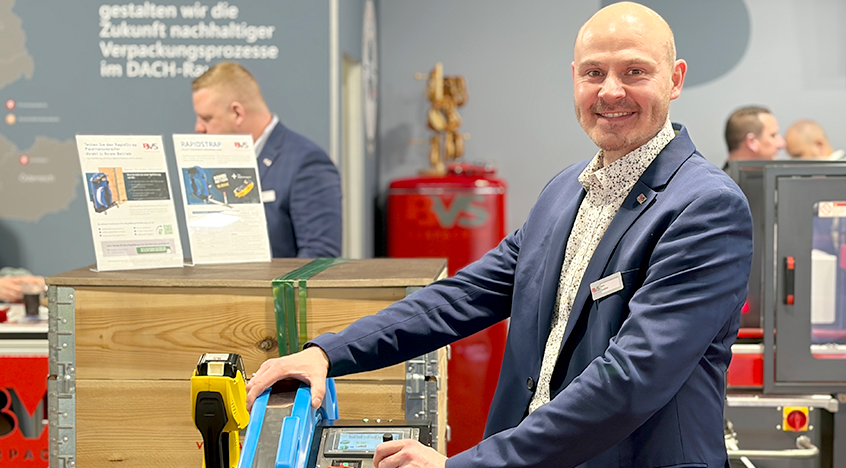 Jens Seldner demonstriert eine Verpackungsanlage und hält ein das Band von Rapidstrap in der Hand