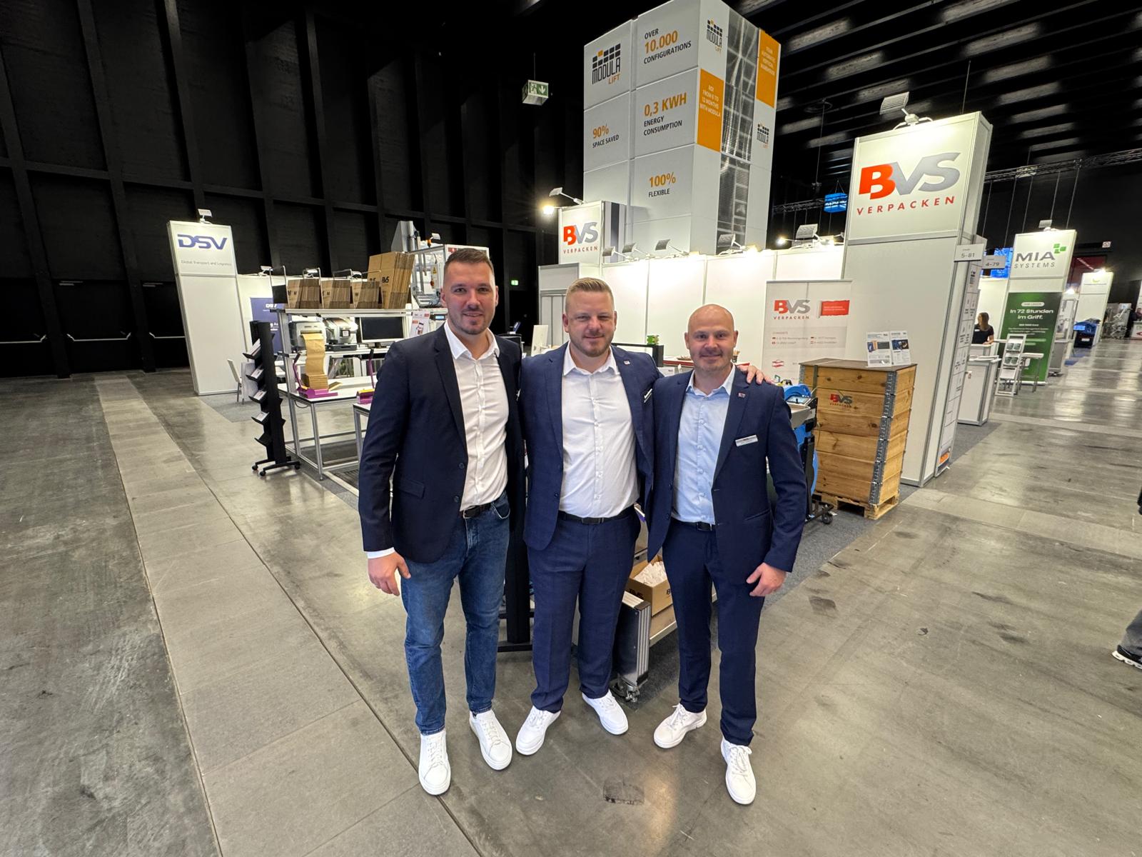 Drei Personen stehen vor dem Messestand von BVS Verpacken in einer Messehalle. Im Hintergrund sind Verpackungslösungen, Maschinen und weitere Ausstellerstände zu sehen.